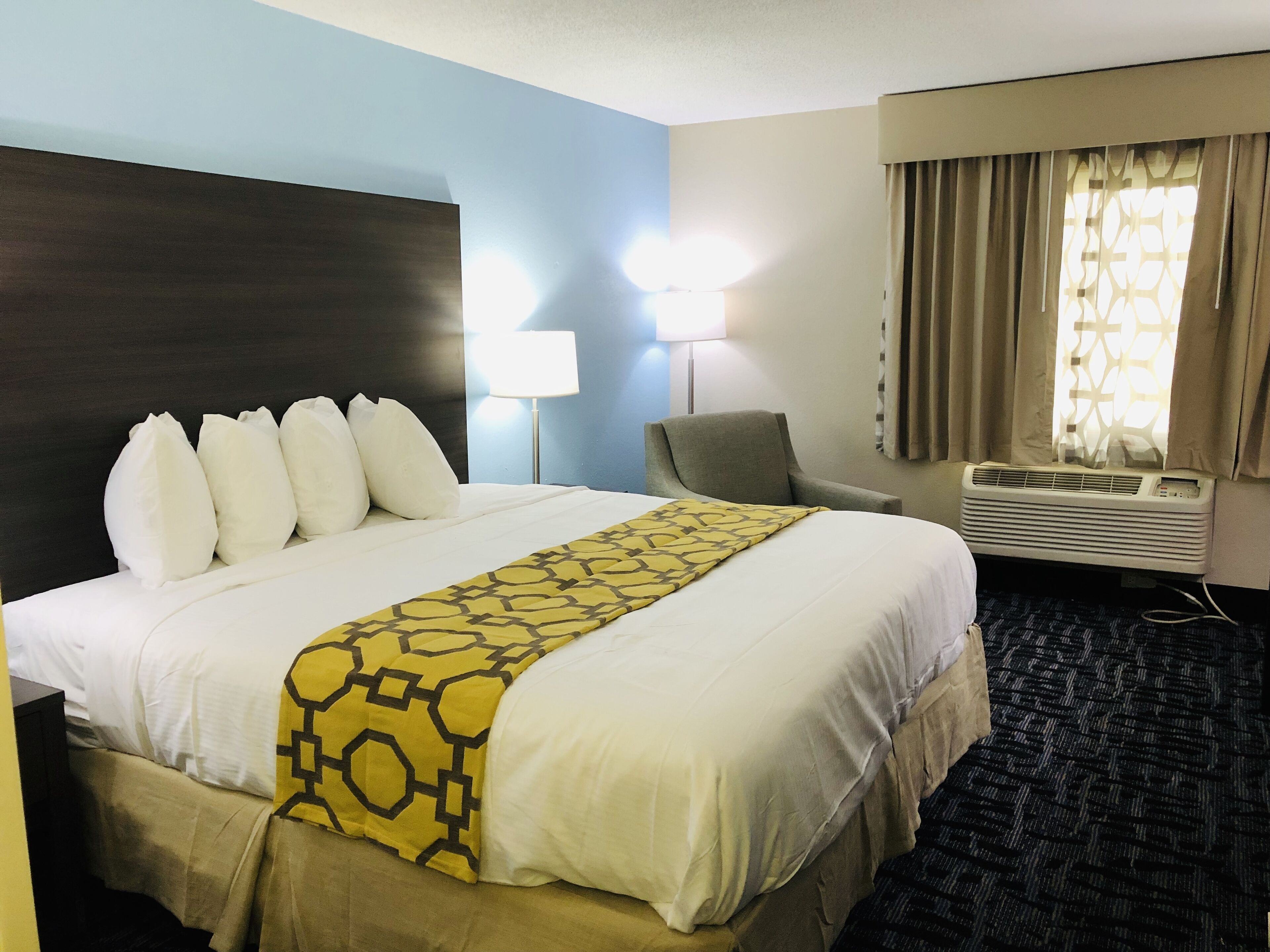 A hotel room with a large bed, two bedside lamps, a chair, an air conditioning unit, and a window with curtains.