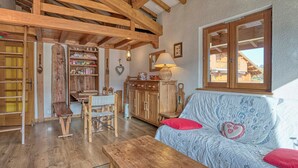 A rustic interior with exposed wooden beams, a wooden dining table, a sofa with red cushions, and a window overlooking a building. - Cozy apartment with mezzanine in Megève, near ski slopes (Megève)