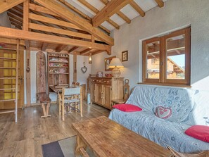 Interior rústico com vigas de madeira aparentes, uma mesa de jantar de madeira, um sofá com almofadas vermelhas e uma janela com vista para um edifício.