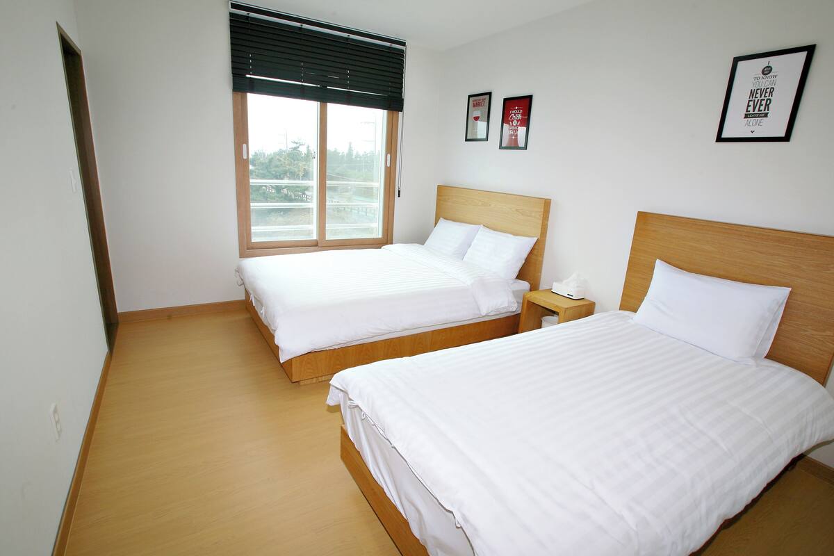 A hotel room with two single beds, a wooden floor, a large window with blinds, and two framed motivational posters on the wall.
