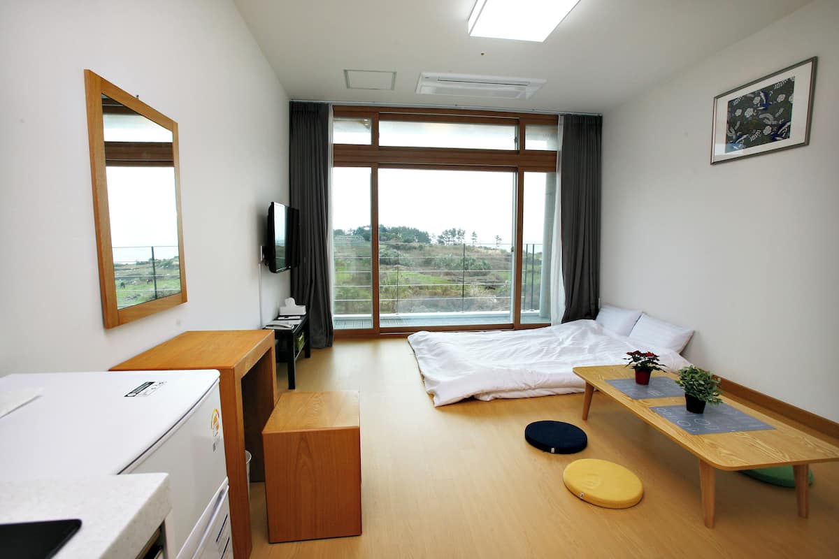 A modern hotel room with a bed, a desk, a small table with plants, a TV, and a large window with a view of the outdoors.