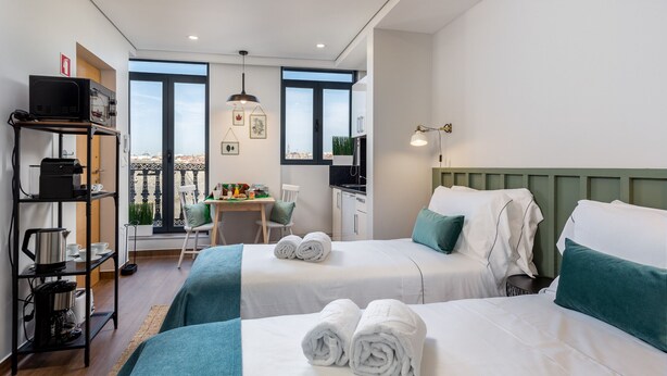 Ein Hotelzimmer mit zwei Betten, einem Schreibtisch, einem Stuhl, einem Regal mit Küchenutensilien und Blick auf die Stadt.