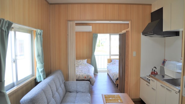 A compact room with a sofa, two beds, a kitchenette, and a window with curtains. - Reiwa House (Ishigaki)