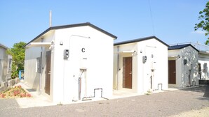 A row of small, white, single-story buildings with numbered doors, likely residential units or small apartments, with a clear sky above. - Reiwa House (Ishigaki)
