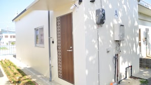 A small, single-story building with a wooden door and a window. There are outdoor electrical boxes and pipes on the exterior wall. - Reiwa House (Ishigaki)