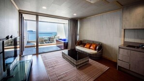 A modern living room with a glass coffee table, a sofa with cushions, a kitchen area, and a view of the sea through large glass doors. - Visby Jeju Palm Village (Seogwipo)