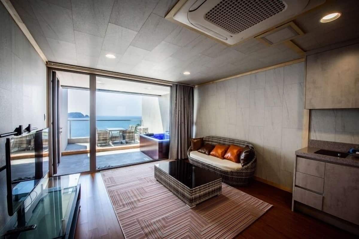 A modern living room with a glass coffee table, a sofa with cushions, a kitchen area, and a view of the sea through large glass doors.