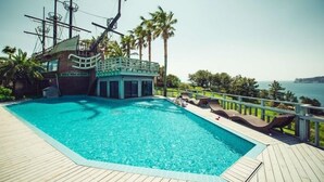 A swimming pool with a wooden deck, surrounded by palm trees and a view of the sea. - Visby Jeju Palm Village (Seogwipo)