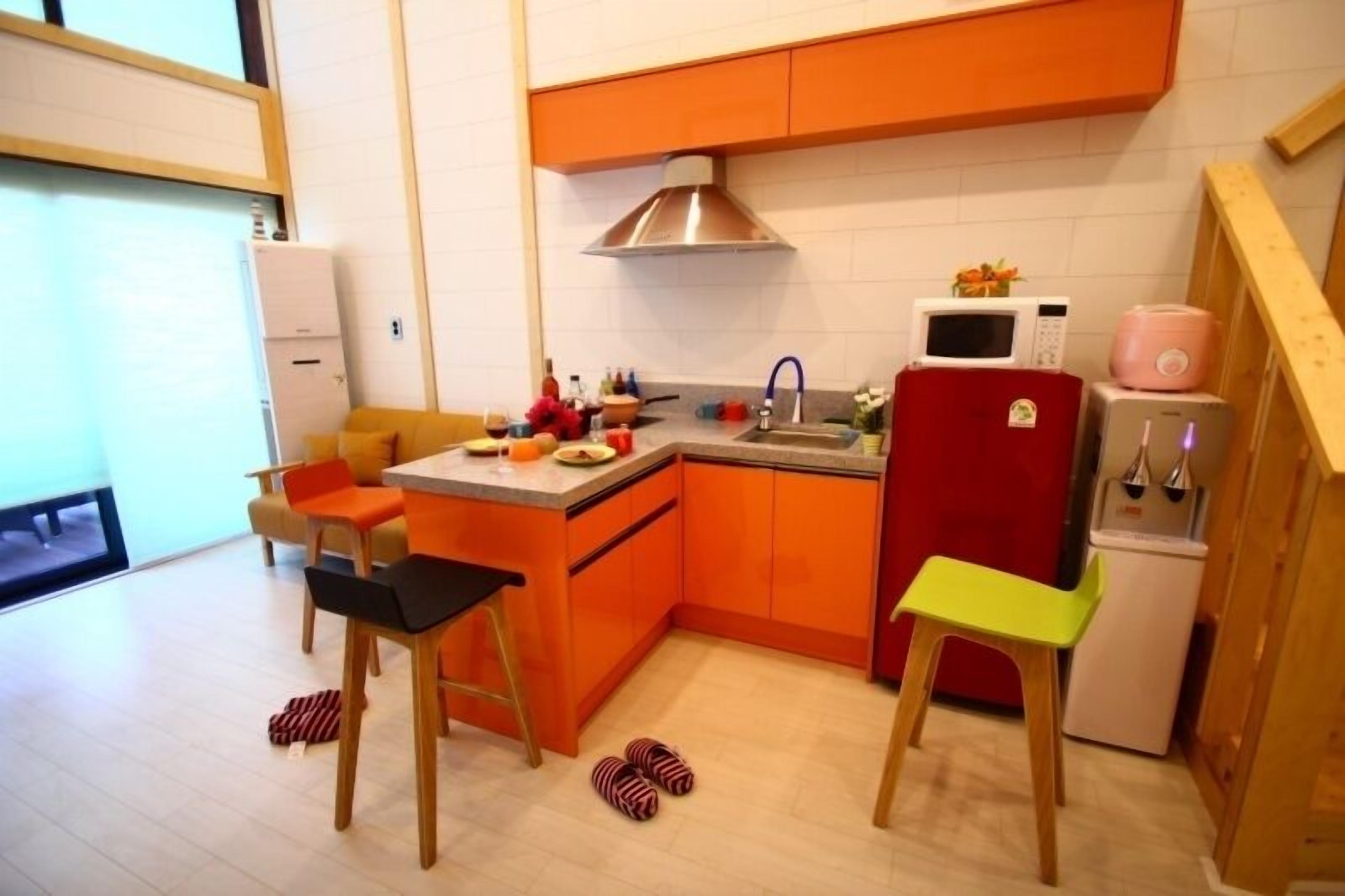 A compact kitchen with orange cabinets, a stainless steel range hood, a microwave, a refrigerator, and a small dining area with mismatched chairs.