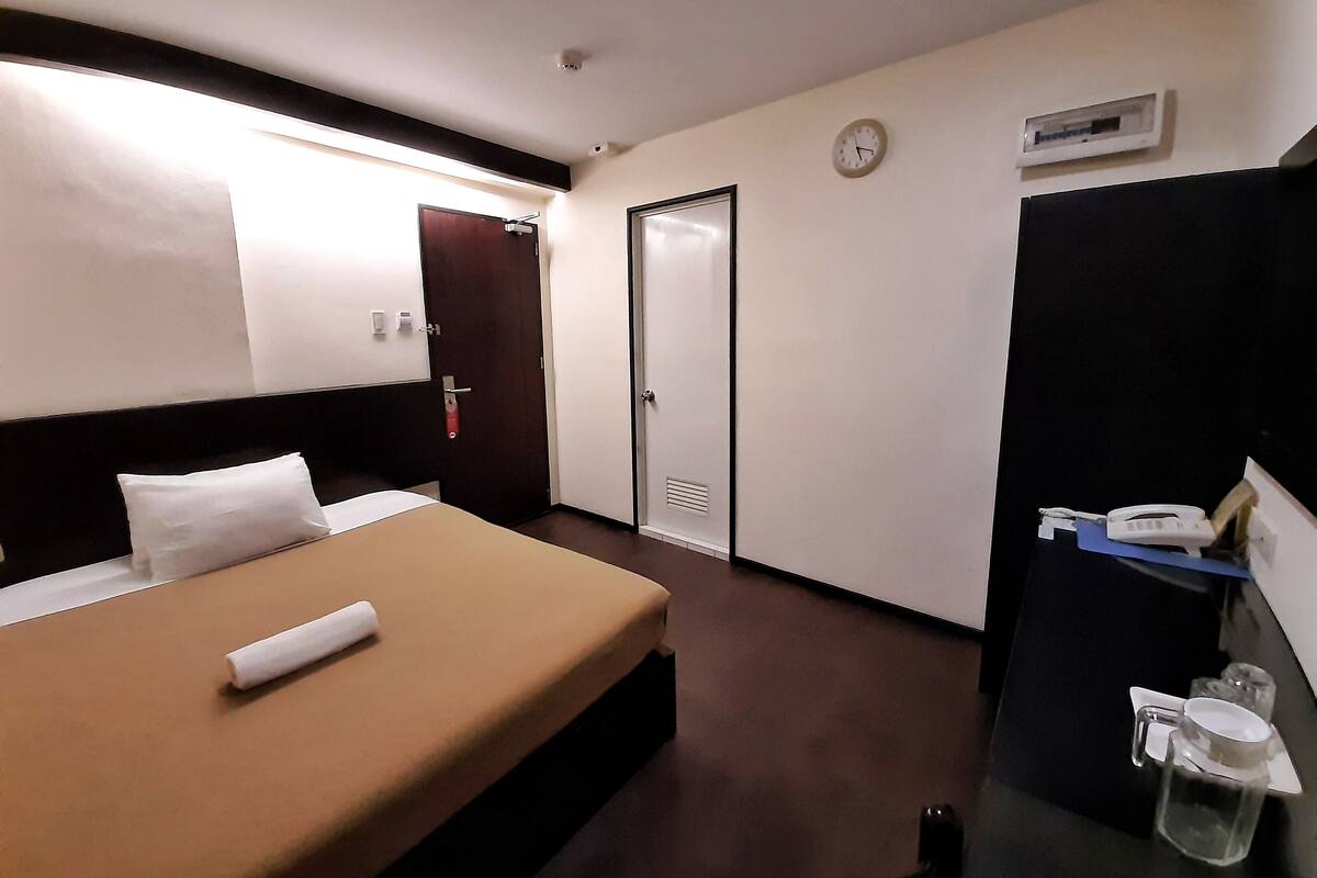 A hotel room with a bed, a bedside table with a telephone and glasses, a door, and a clock on the wall.