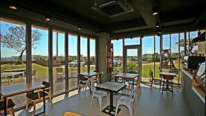 A spacious indoor area with large windows offering a view of the outdoors. There are several tables and chairs arranged for dining or seating. - Villanoi Jeju (Seogwipo)