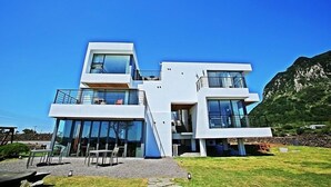 A modern two-story house with large glass windows and a balcony, set against a mountainous backdrop. - Villanoi Jeju (Seogwipo)
