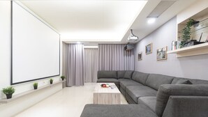 Ein modernes Wohnzimmer mit einem grauen Ecksofa, einem Couchtisch und einem großen Flachbildfernseher. Auf einem Regal stehen Blumentöpfe und an der Decke ist ein Projektor angebracht.