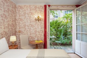 A room with a bed, a chair, a table with a lamp, and a view of a garden through an open door. - Hôtel des Grandes Ecoles (Paris)