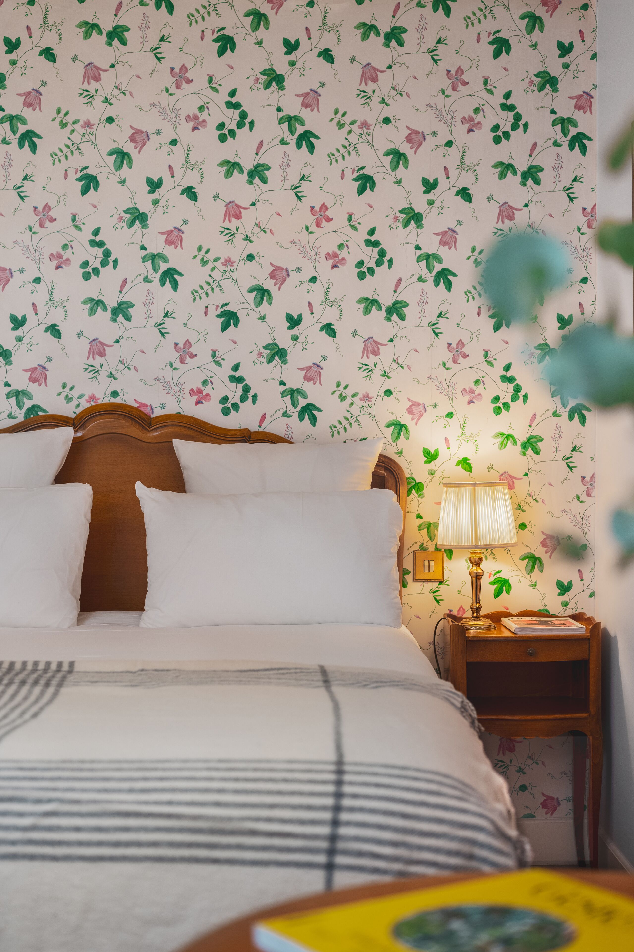 A neatly made bed with white pillows and a striped blanket, a wooden headboard, a bedside lamp, and a floral wallpaper pattern.