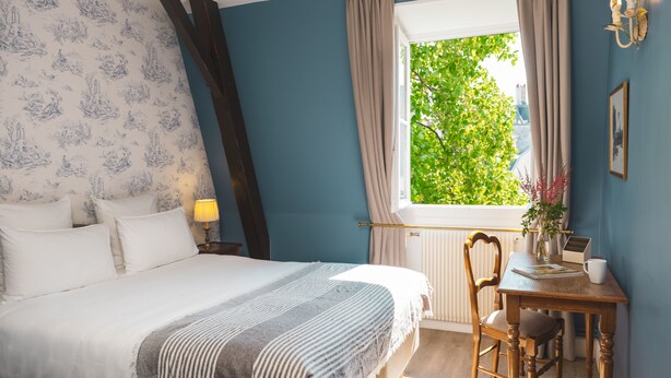 A bedroom with a bed, a window with curtains, a desk with a chair, and a wall-mounted lamp.