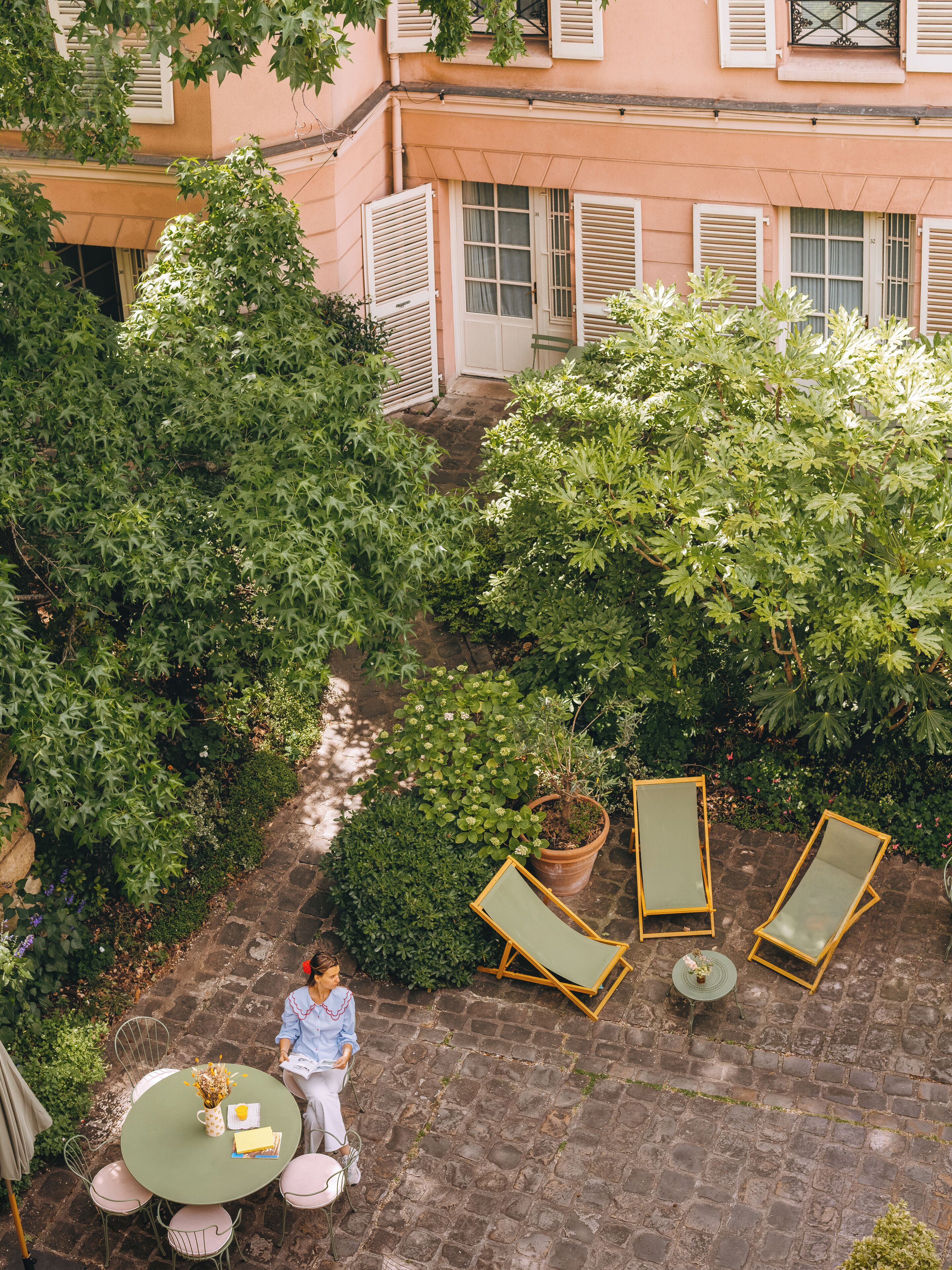A courtyard with a table and chairs, surrounded by greenery and a building with shutters.
