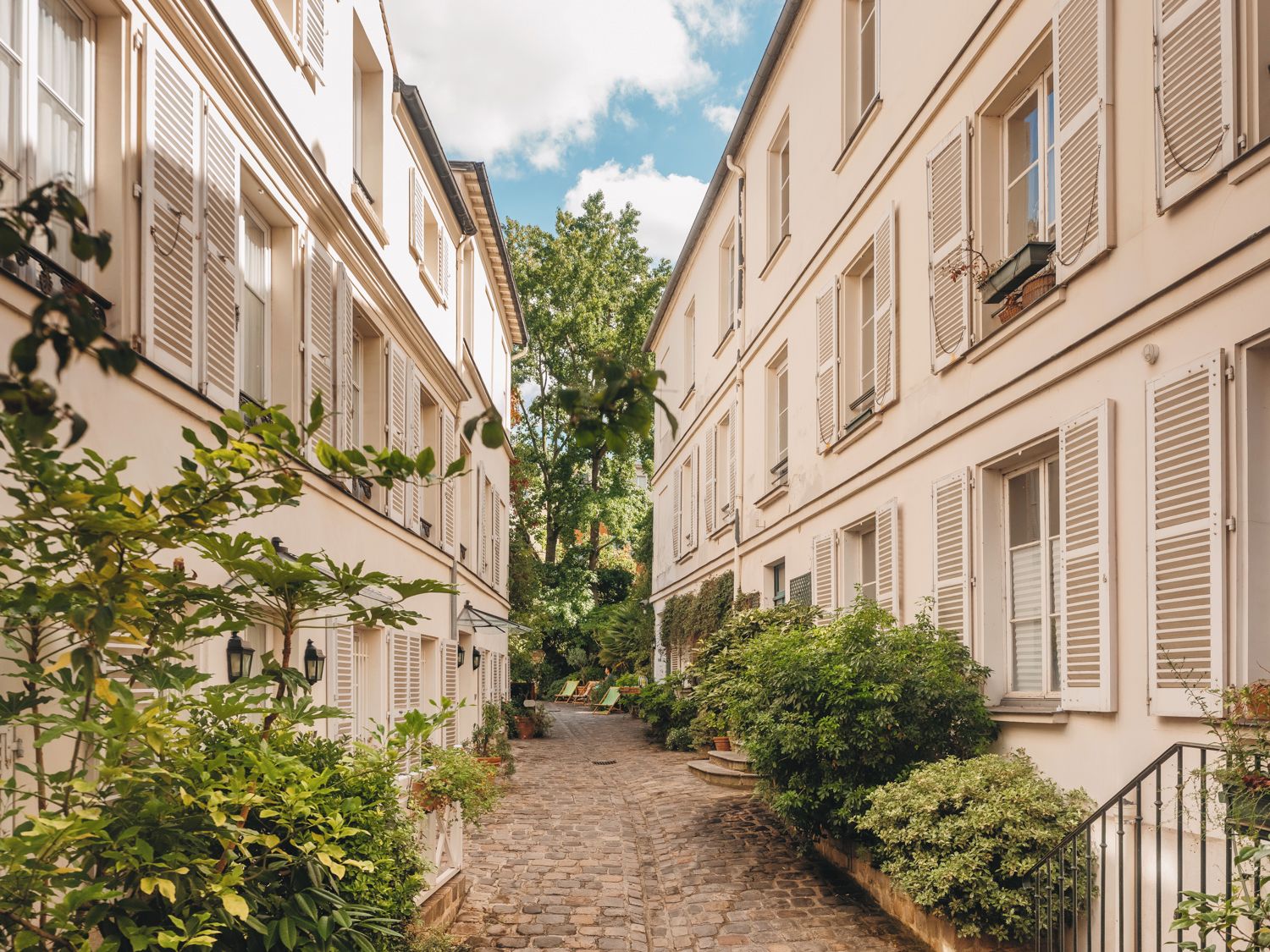 A narrow cobblestone street flanked by buildings with shutters, greenery, and a clear sky.