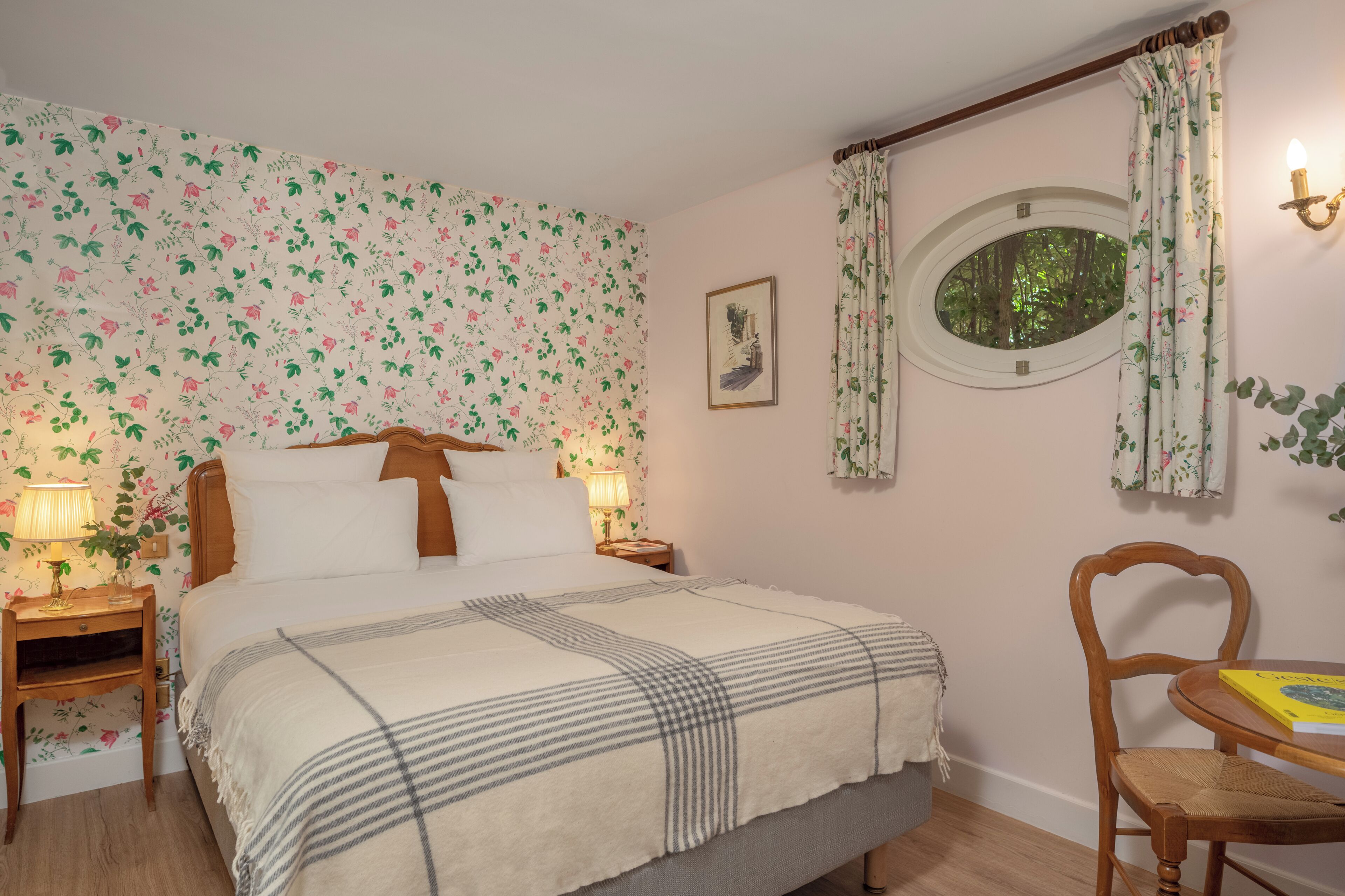 Un dormitorio con papel tapiz floral, una cama con manta de cuadros, dos lámparas de noche, un cuadro en la pared y una ventana con vistas a árboles.
