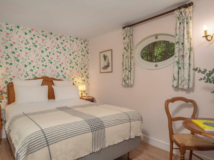 Un dormitorio con papel tapiz floral, una cama con manta de cuadros, dos lámparas de noche, un cuadro en la pared y una ventana con vistas a árboles.