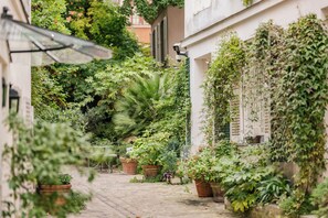 A narrow cobblestone path flanked by lush greenery and potted plants, leading to a building with a white facade and a security camera. - Hôtel des Grandes Ecoles (Paris)