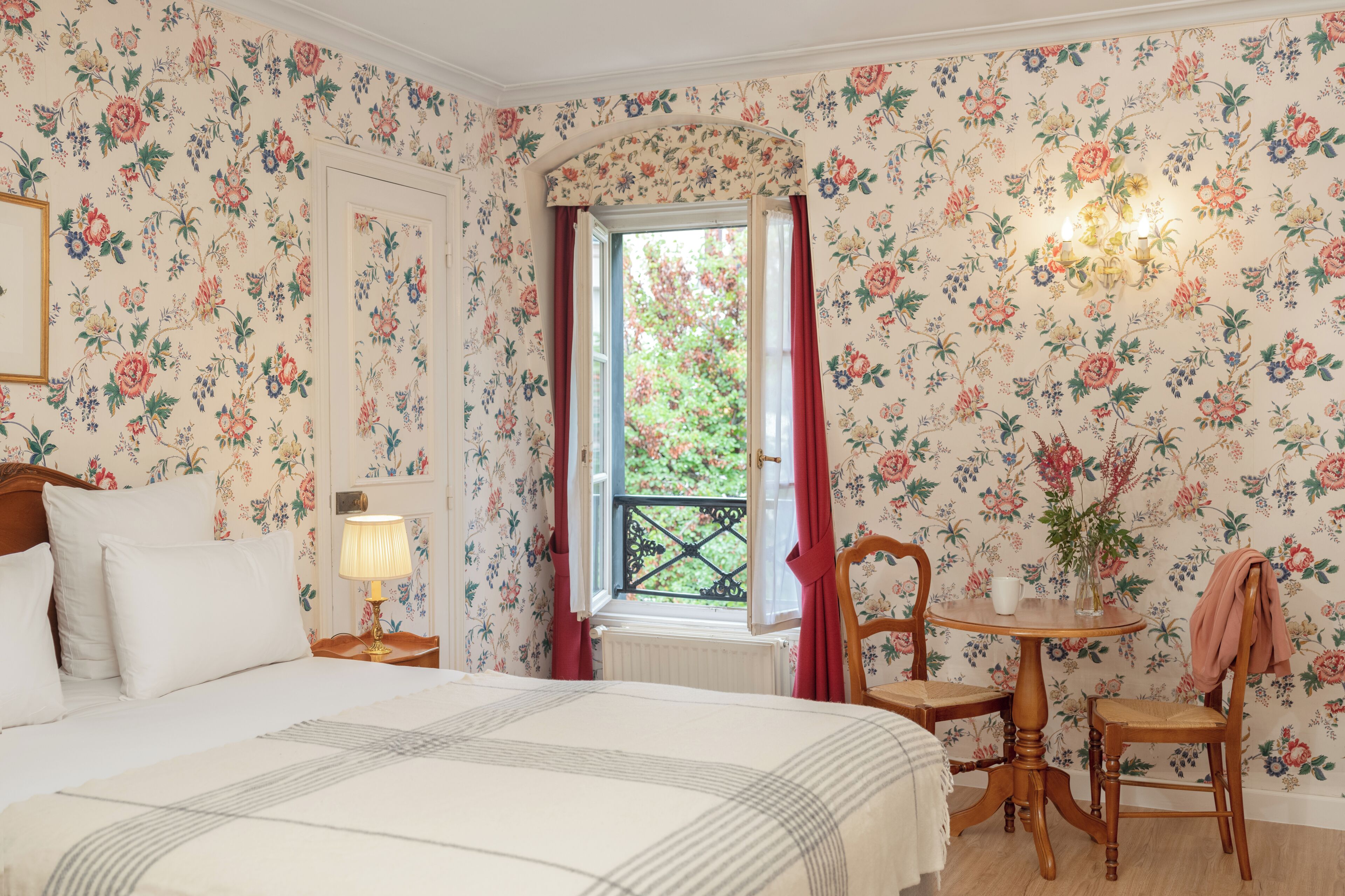 A bedroom with floral wallpaper, a bed with white linens, a nightstand with a lamp, a window with red curtains, and a small table with a chair.