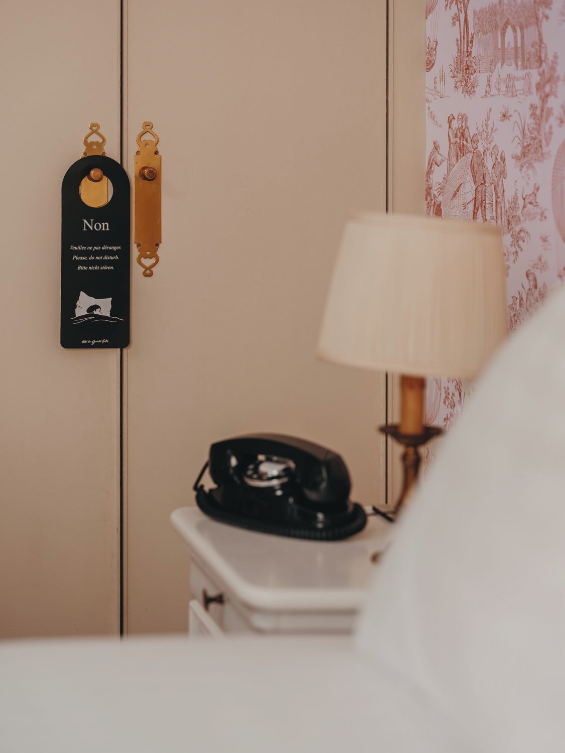 A hotel room with a bedside telephone, a lamp, and a wall-mounted sign with a message in a foreign language.