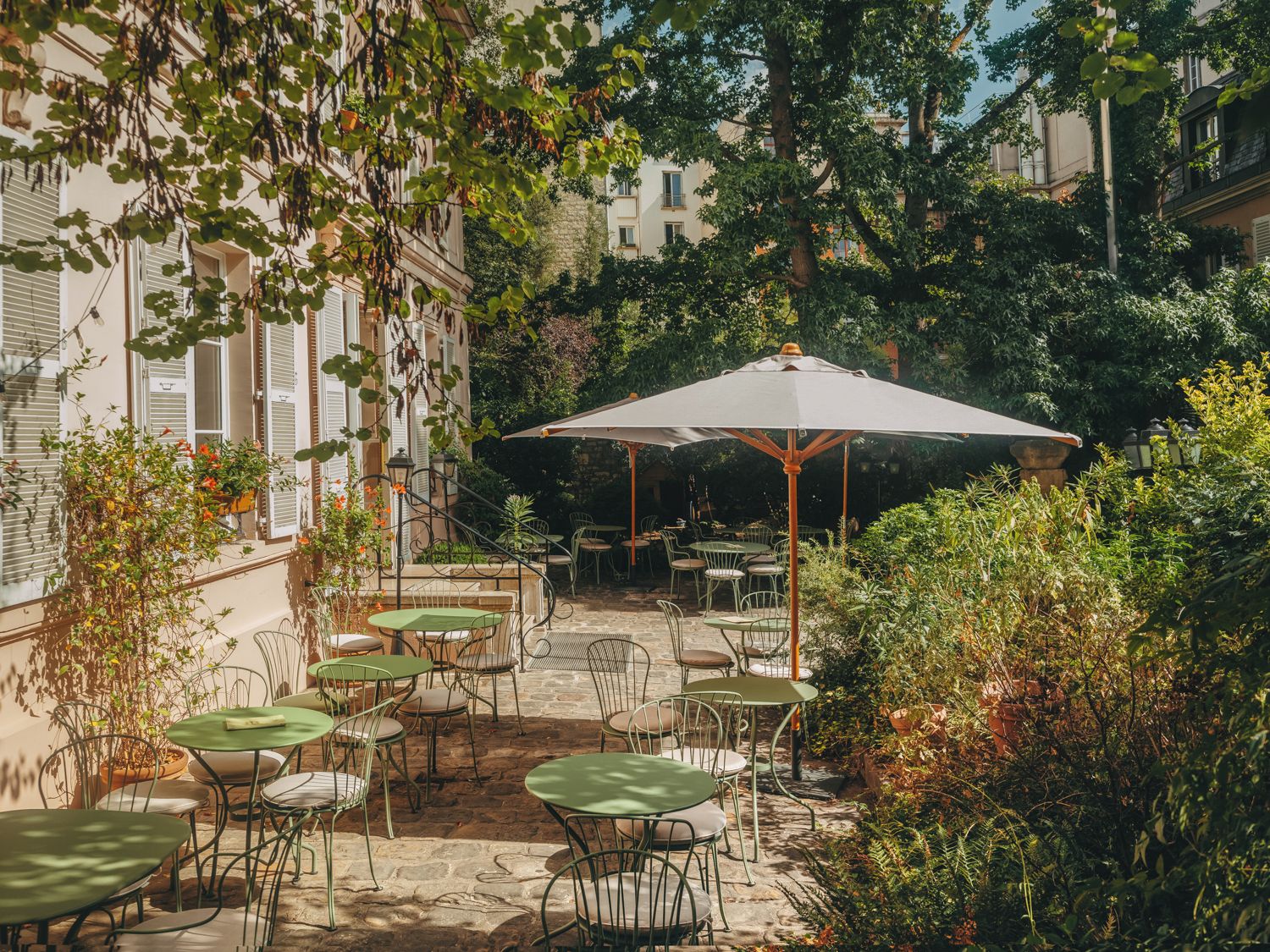 An outdoor patio area with tables and chairs, surrounded by greenery and buildings.