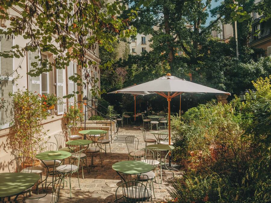 An outdoor patio area with tables and chairs, surrounded by greenery and buildings.