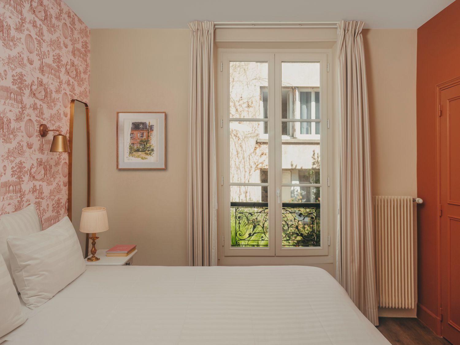 A bedroom with a bed, a nightstand, a lamp, a framed picture, a window with curtains, and a radiator.