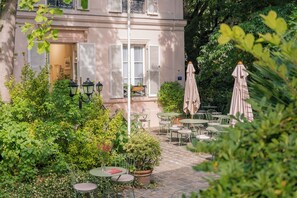 An outdoor patio area with tables and chairs, surrounded by greenery and a building with shutters. - Hôtel des Grandes Ecoles (Paris)