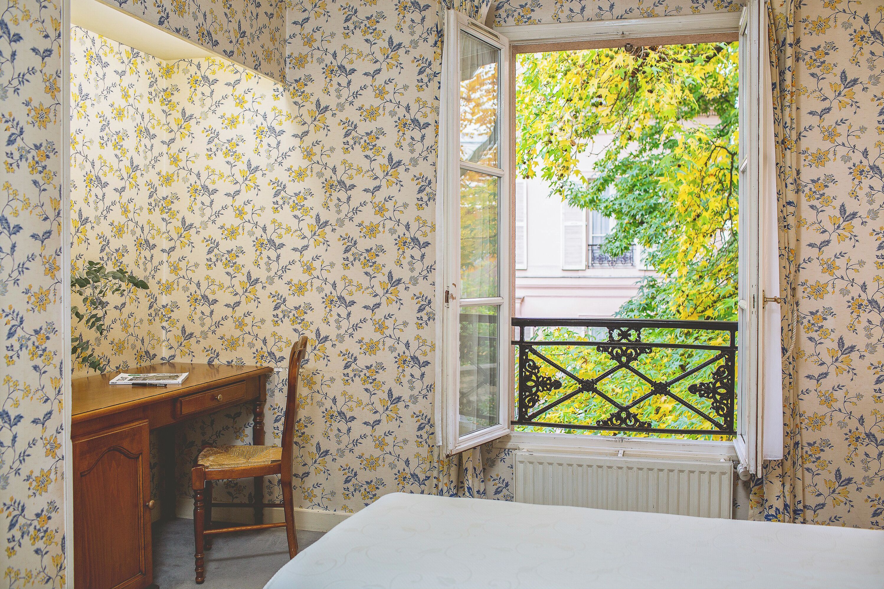 A room with a bed, a wooden desk, a chair, and a window with a view of greenery.
