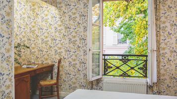 A room with a bed, a wooden desk, a chair, and a window with a view of greenery.