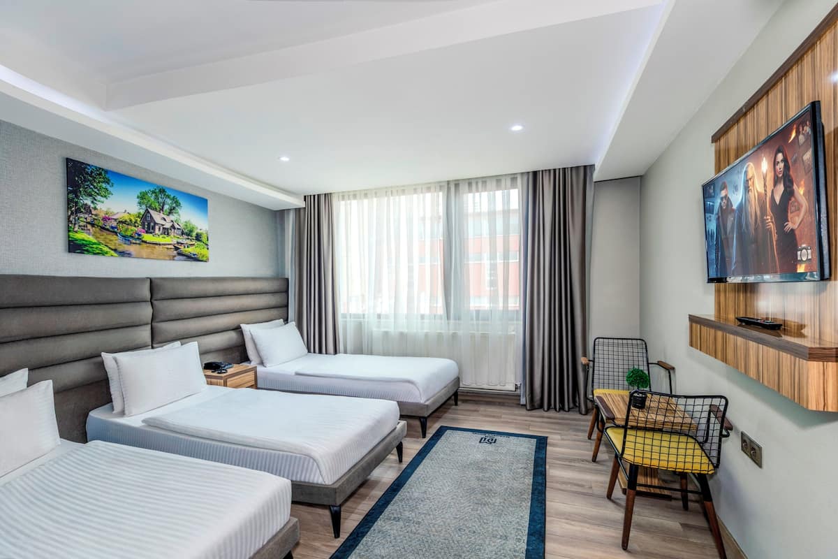 Quarto de hotel com duas camas, uma televisão, um quadro, área de refeições com mesa e cadeiras, e janela com cortinas.
