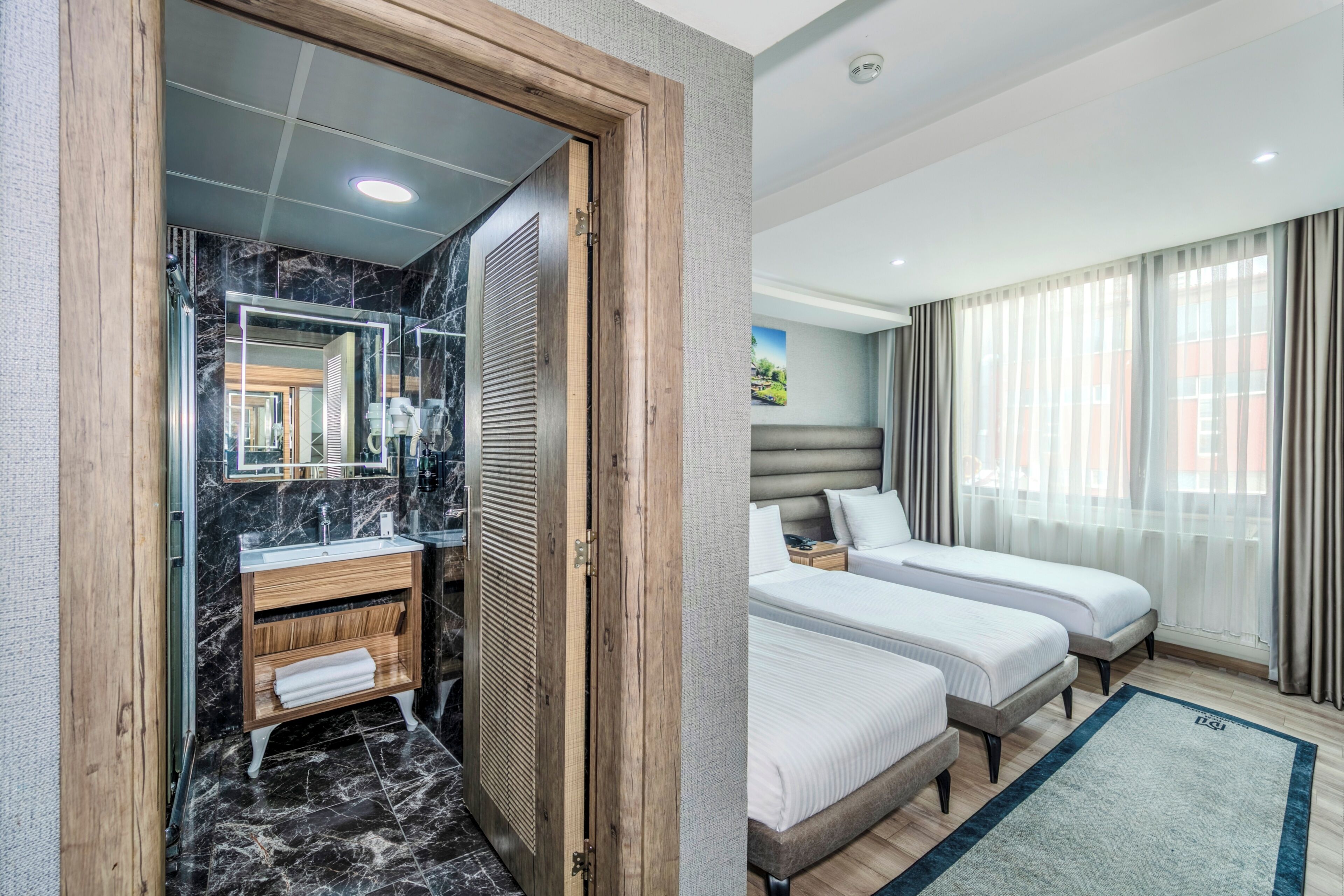 A hotel room with a bathroom, two beds, a nightstand, and a large window with curtains.