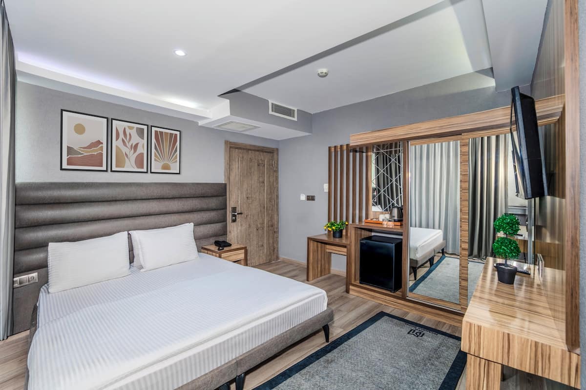 Quarto de hotel moderno com uma cama grande, cabeceira de madeira, televisão, espelho e uma pequena planta.