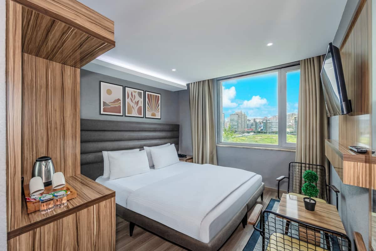 Quarto de hotel moderno com uma cama grande, mesa de cabeceira de madeira com cafeteira, pequena mesa com planta e vista para a cidade pela janela.