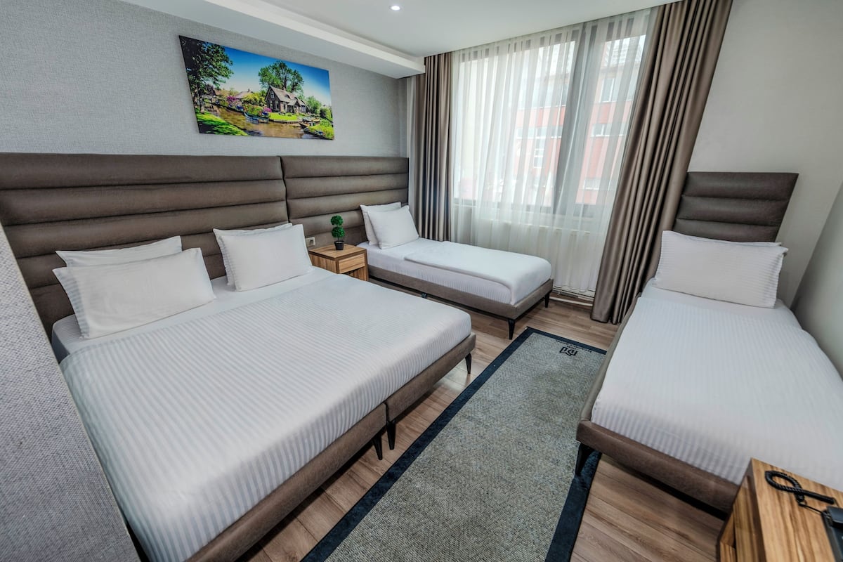 Quarto de hotel com duas camas, uma pequena mesa de cabeceira com uma planta, um quadro na parede e vista para a cidade pela janela.