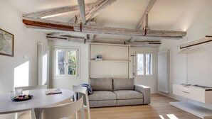 A modern living room with exposed wooden beams, a grey sofa, a white dining table with chairs, and a white cabinet. - Venice Skyline View Sudio Apartment (Venice)
