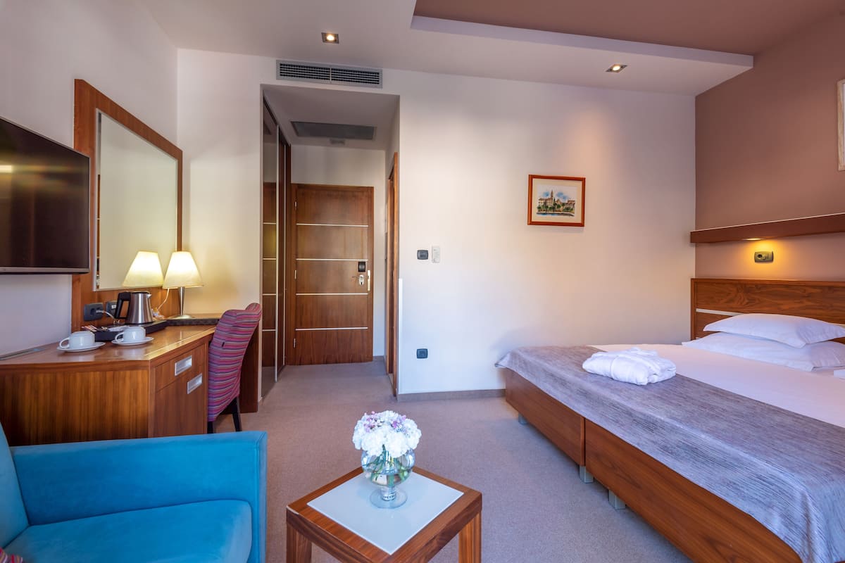 Ein Hotelzimmer mit einem Bett, einem Schreibtisch mit Lampe, einem Sessel, einem kleinen Tisch mit Vase und einem Bild an der Wand.