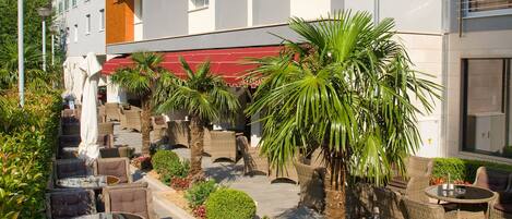 Un hotel con terraza al aire libre, palmeras y montañas al fondo.