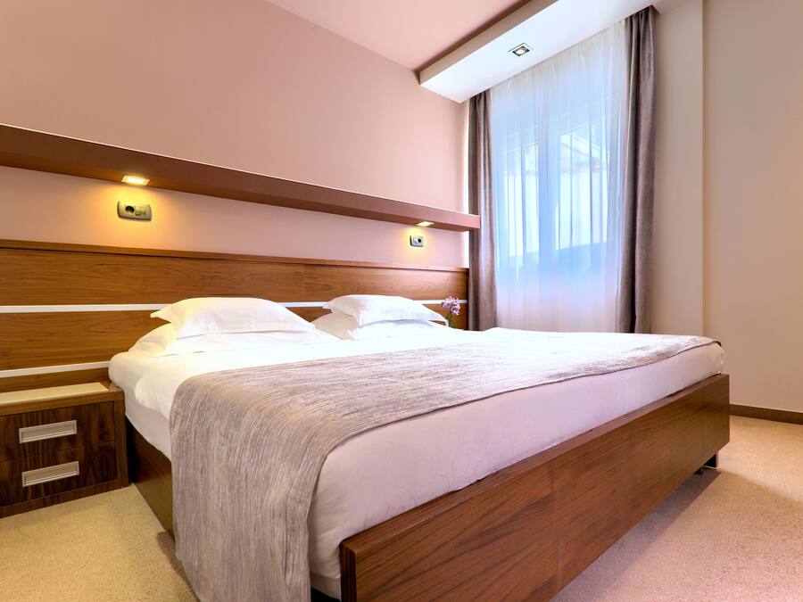 A neatly made bed with a wooden headboard, a bedside table with drawers, and a window with curtains.