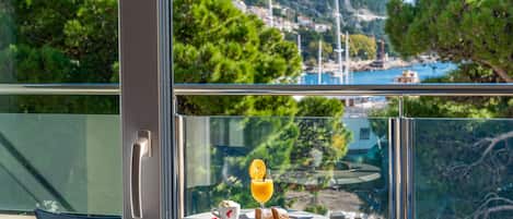 Un balcón con una mesa lista para el desayuno, con vistas a una zona costera con árboles y edificios.