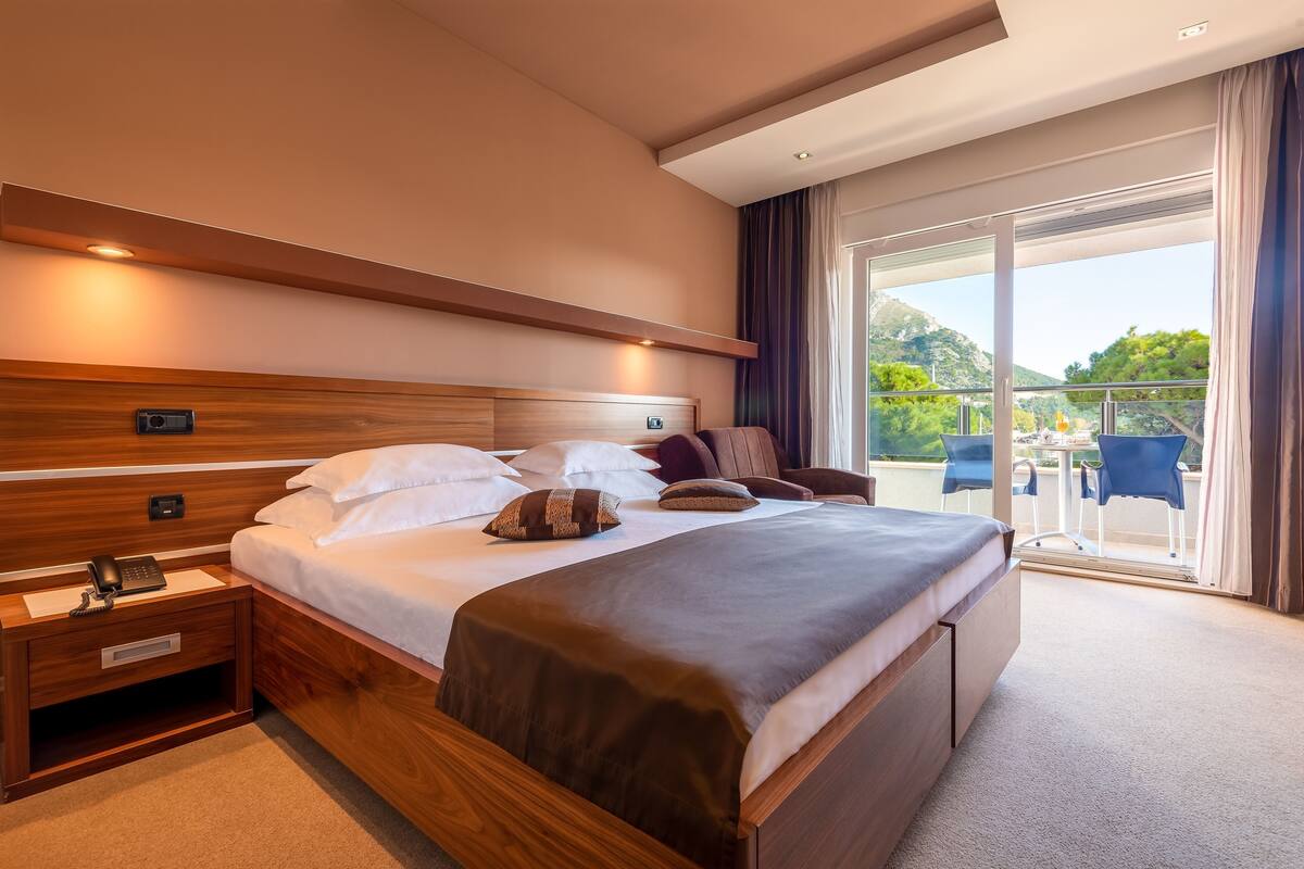 Ein Hotelzimmer mit einem großen Bett, Nachttischen, einer Bedienfeld und einem Balkon mit Sitzgelegenheit.