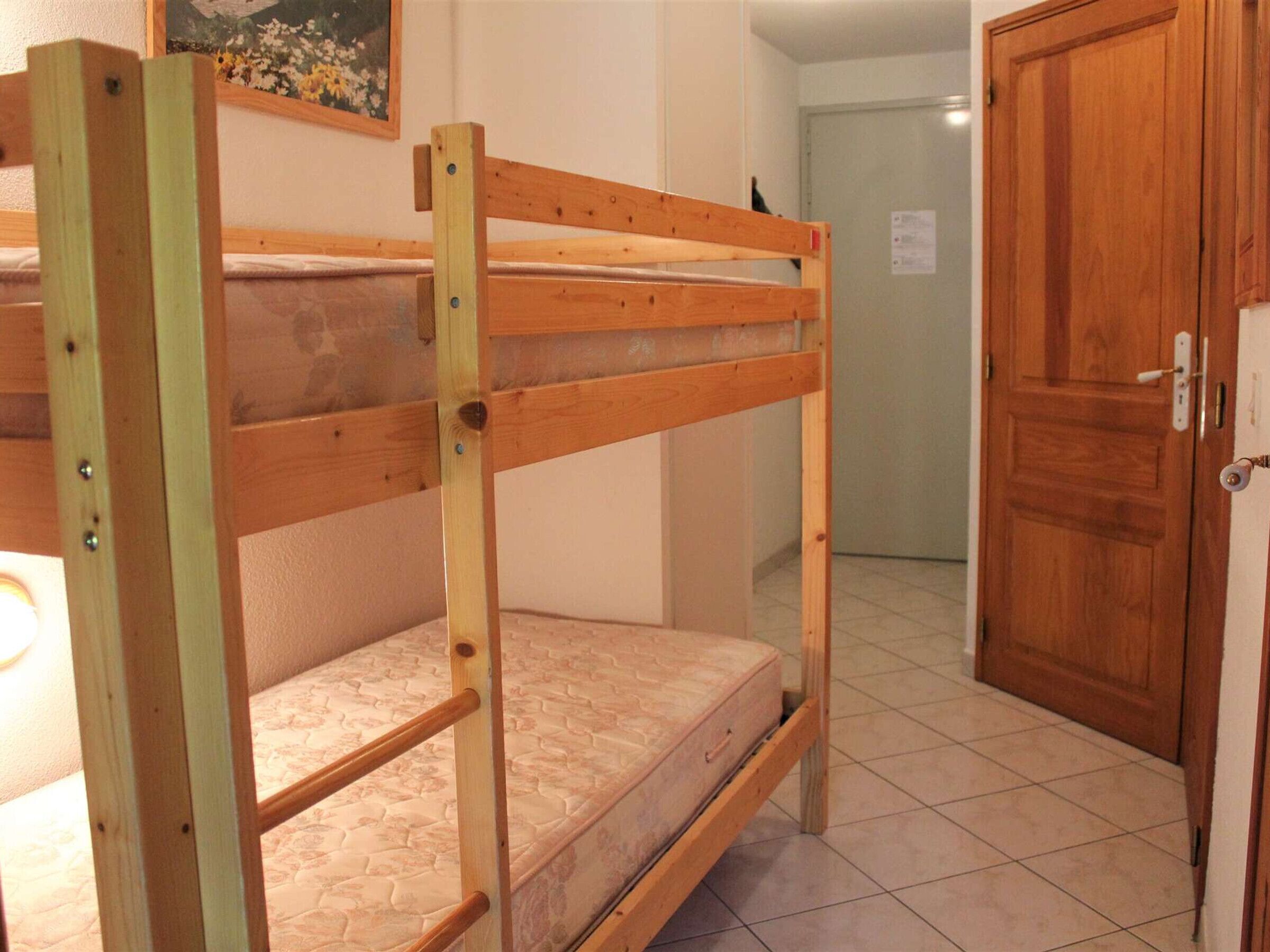 Una cama individual en literas en una habitación con puerta de madera y suelo de baldosas.