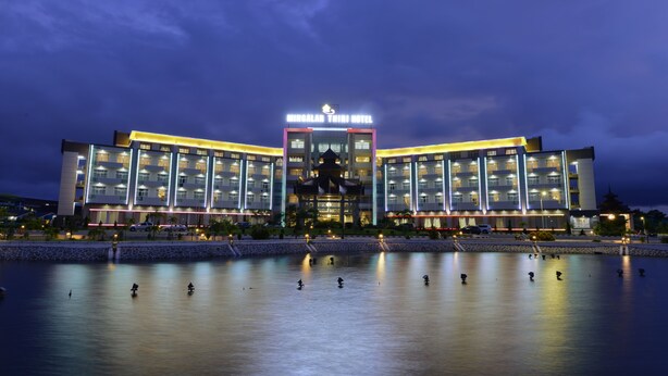 Un hotel moderno con entrada central y varios pisos, rodeado por una masa de agua tranquila que refleja las luces.