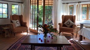 A room with a thatched roof, two armchairs with cushions, a wooden table with a vase of flowers, and a bed with a white bedspread. - Waterberg Cottages (Vaalwater)
