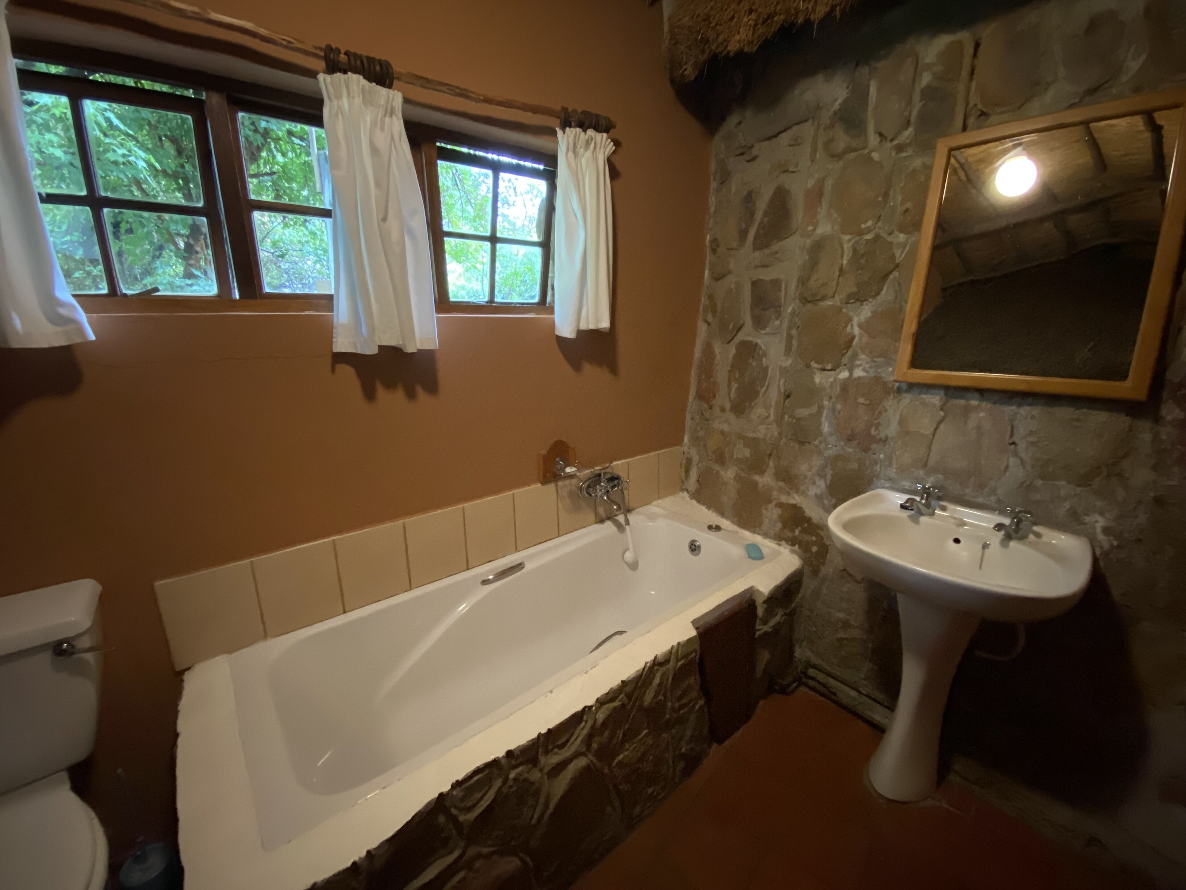 A bathroom with a stone wall, a bathtub, a sink, and a window with curtains.