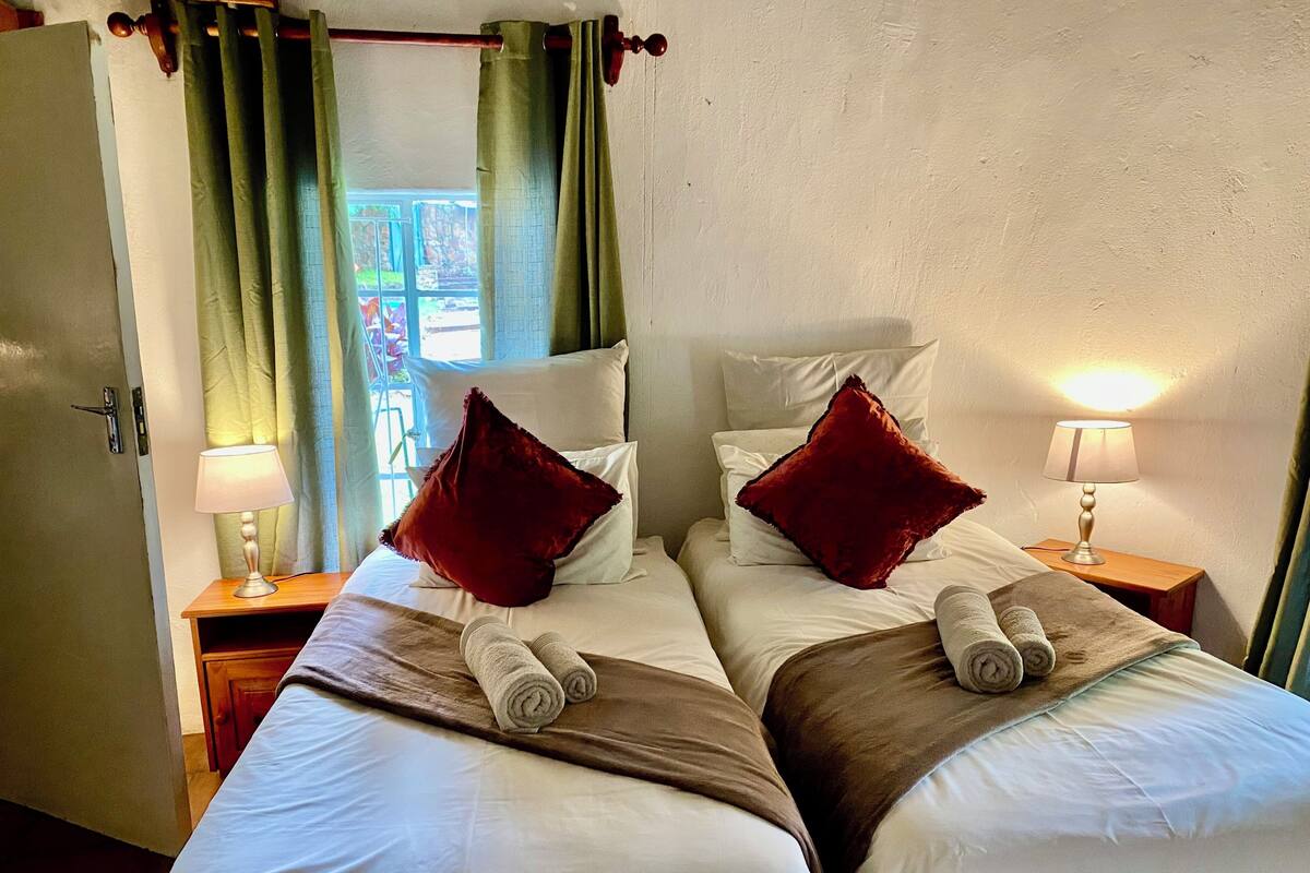 A hotel room with two beds, a door, a window with curtains, and a nightstand with a lamp.
