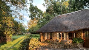 A rustic stone cottage with a thatched roof, surrounded by lush greenery and a well-maintained lawn. - Waterberg Cottages (Vaalwater)